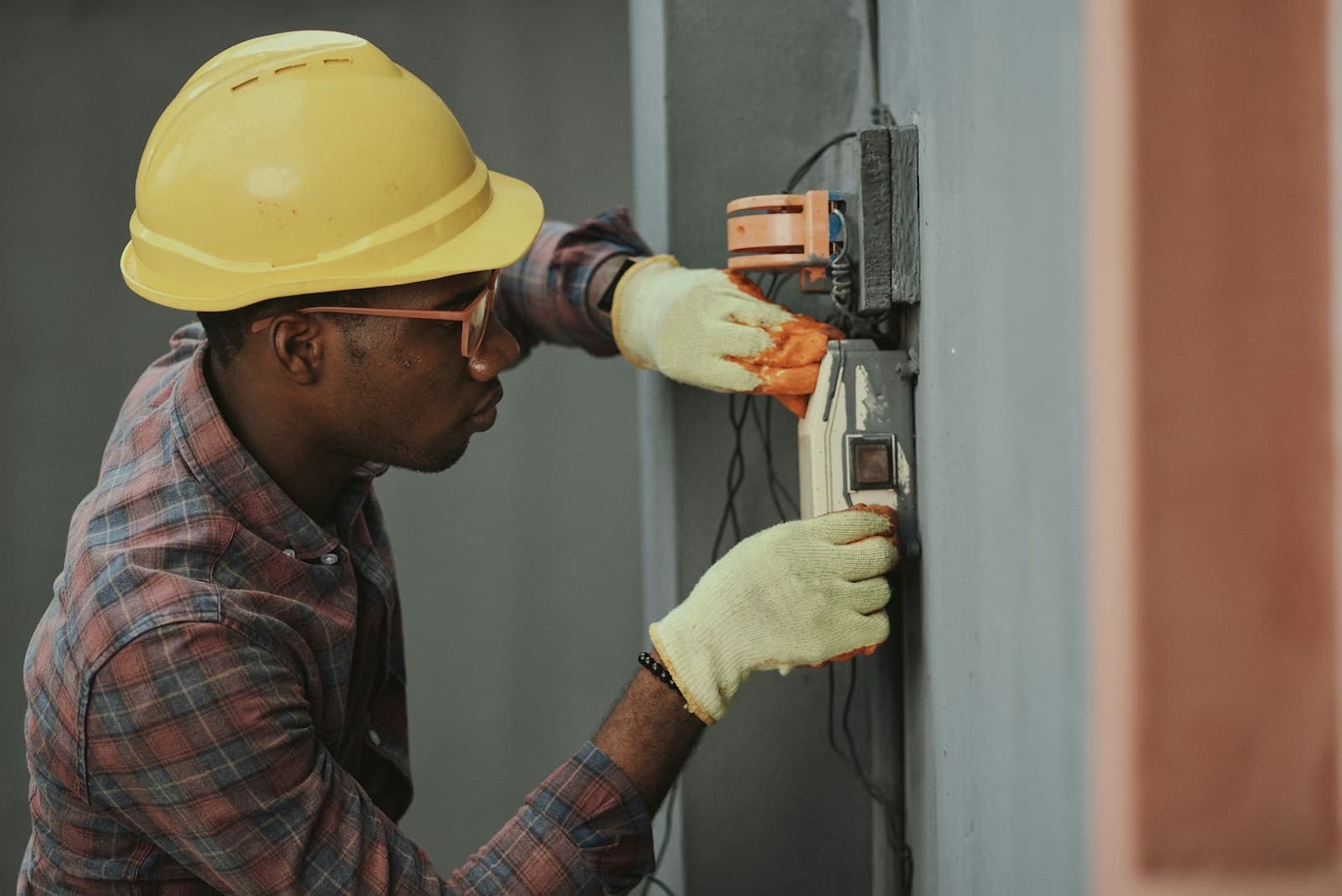 An electrician in a yellow hard hat and work gloves fitting wiring into a box on a site wall